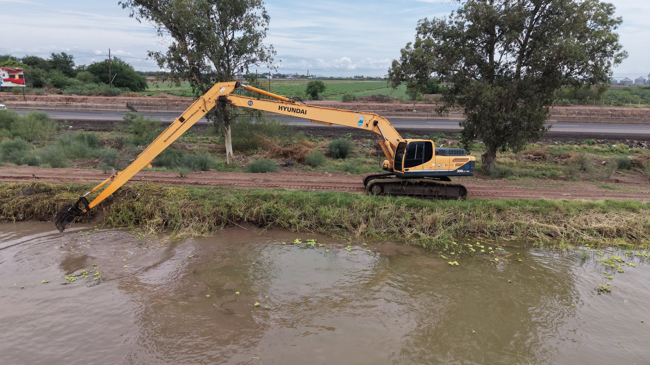 Limpieza de canal con brazo largo. Excavadora trabajando sobre canal con agua y retiro de vegetación.